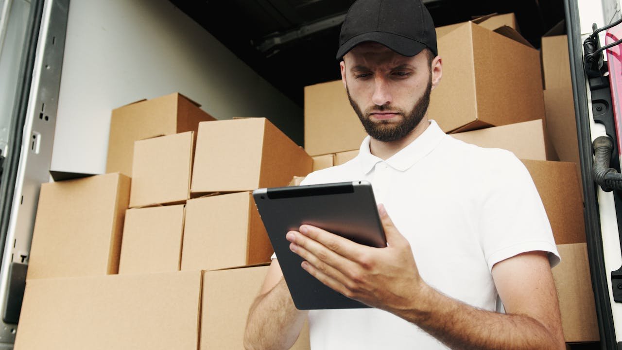 our-story-01 Delivery worker using a tablet to manage shipments with stacked boxes in the background.