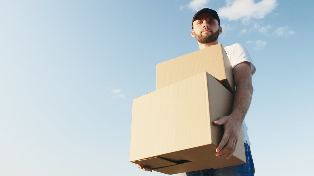portfolio-05 Man in casual attire with beard holding cardboard boxes under a bright blue sky, symbolizing delivery service.