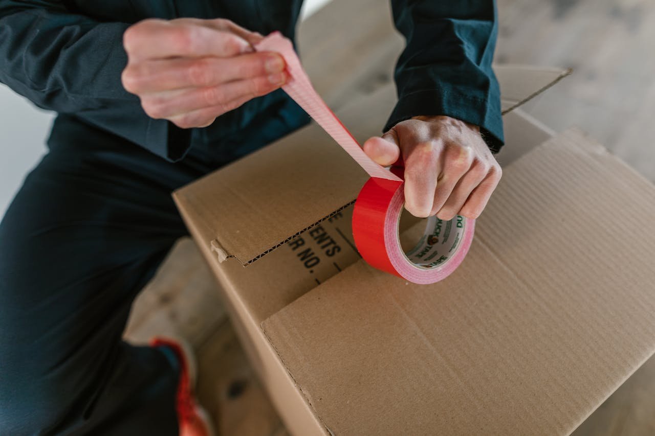 Focus on hands sealing a cardboard box with red tape, depicting careful packaging.